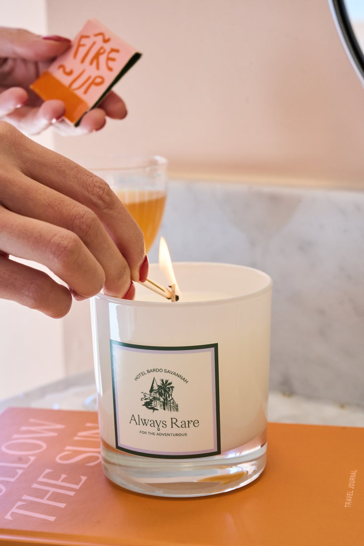 A hand holds a lit match to ignite a white scented candle in a clear glass jar. The candle label reads 'Always Rare'