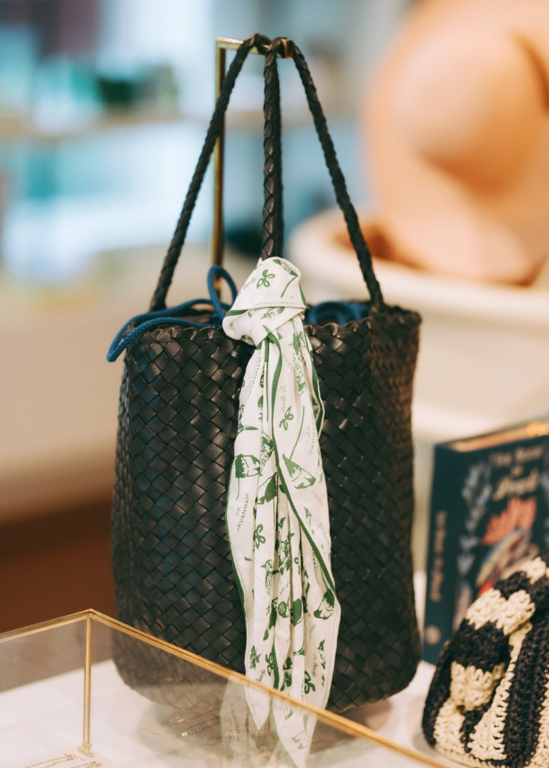 A green woven straw tote bag with braided shoulder straps, styled with a white silk scarf featuring green botanical patterns tied around the handle, displayed on a table in a boutique setting.
