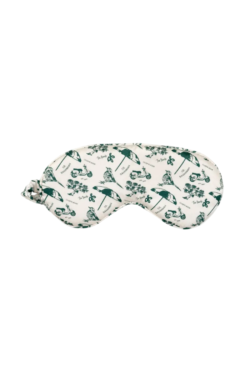 A sleep mask with a dark green savannah pattern featuring umbrellas, birds, and Vespas on an off-white background.