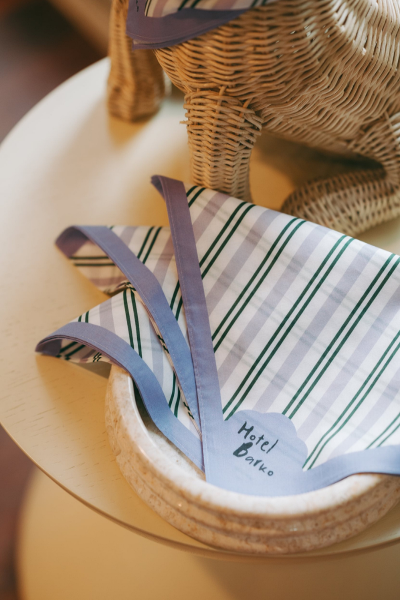 A striped dog scarf with lavender-purple borders, folded in a bowl. The scarf has 'Hotel Barko' in black text on the front.