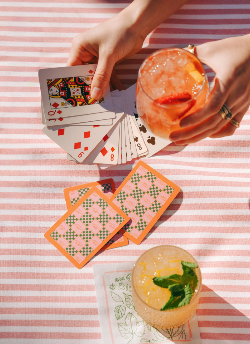 A lifestyle shot of a person's hands holding playing cards and a pink striped cocktail glass on a pink and white striped blanket, with two patterned playing cards and a yellow drink in the foreground.