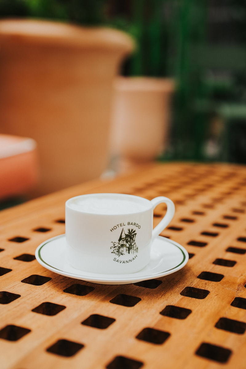 A white ceramic espresso cup with a matching saucer, printed with the Hotel Bardo Savannah logo, set on a wooden table with a blurred outdoor background.