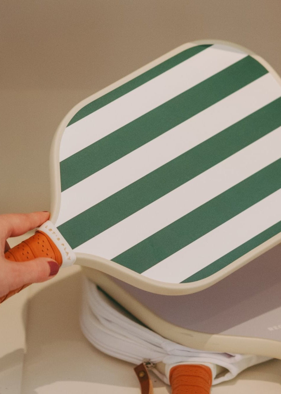 A hand holds a white paddle with green diagonal stripes and an orange wrapped handle on a shelf, with two more paddles stacked below and an orange striped product box to the right.