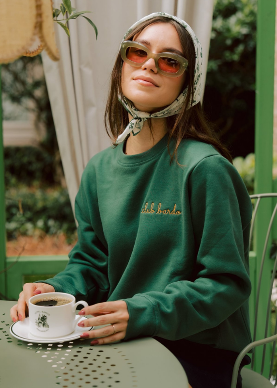 A woman wearing a dark green crewneck sweatshirt with gold 'club bardo' embroidery sits at an outdoor patio table holding a cup of coffee. She is wearing green sunglasses and patterned headscarf.