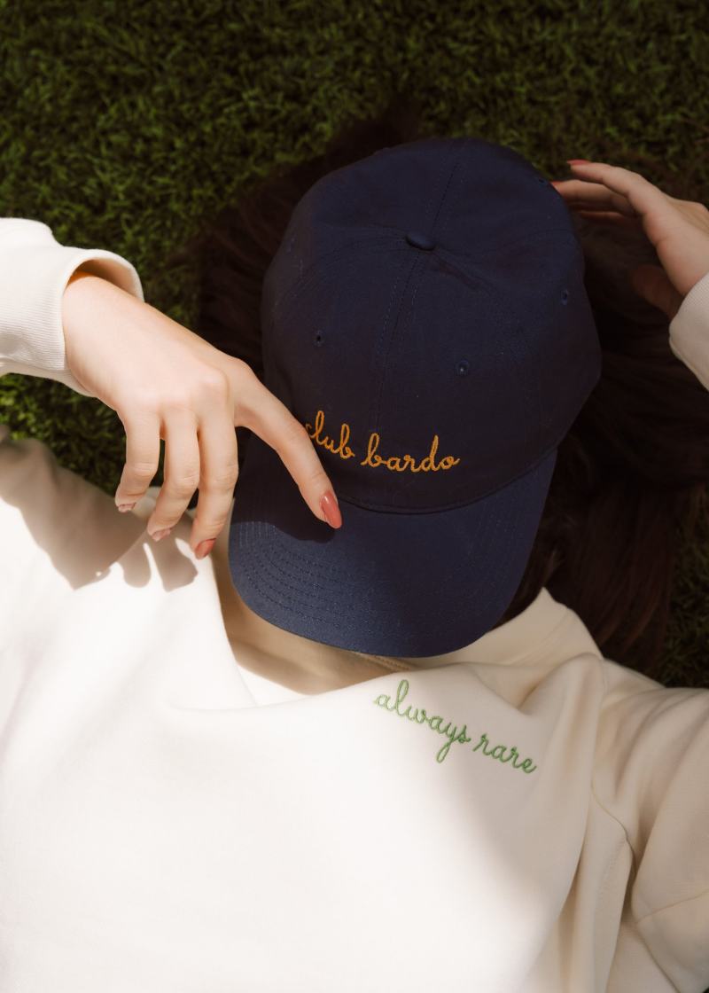A navy blue baseball cap with gold embroidered 'club bardo' script, worn by a person lying on green grass. The person is wearing a cream sweatshirt with green embroidered text.