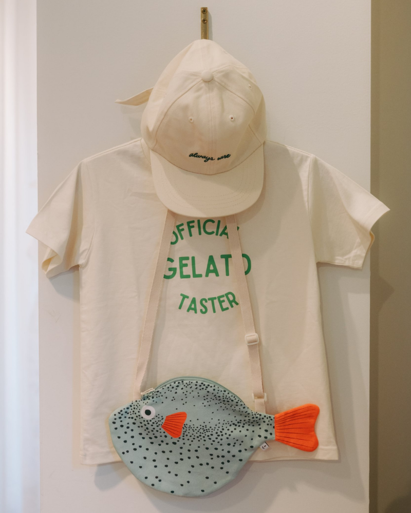 A cream-colored t-shirt with green text, a matching cap, and a fish-shaped crossbody bag with a speckled blue-gray body, orange fin, and green border, hanging on a wall.